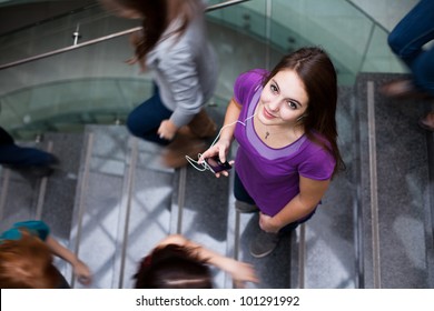 At the university/college - Students rushing up and down a busy stairway - confident pretty young female student looking upwards while listening to music on her mp3 player (color toned image)