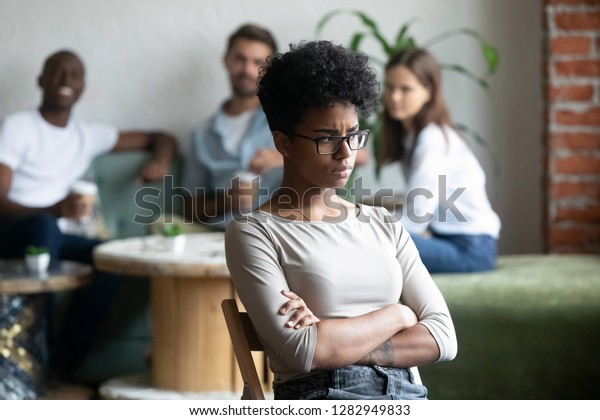 Unkind teenagers looking laughing at black\
classmate, focus on mixed race unhappy shy and frustrated girl\
wearing glasses sitting at closed posture arms crossed feeling\
insecure having low\
self-esteem
