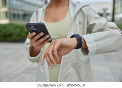 Unknow sportswoman holds digital mobile phone checks fitness tracker results of workout or body vitals on smartwatch dressed in anorak poses outdoors uses modern devices for sport. Cropped shot - Shutterstock ID 2055946859
