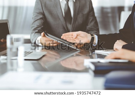 Unknown businessman using tablet computer and working together with his colleague while sits at the glass desk in modern office. Teamwork and partnership concept