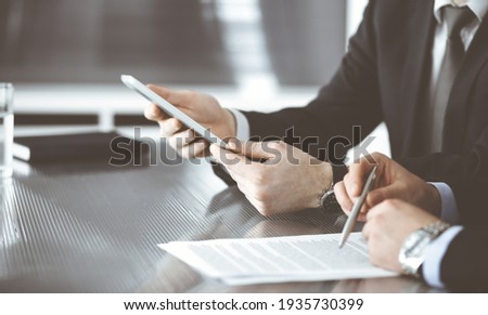 Unknown businessmen using tablet computer and work together at the glass desk in modern office, close-up. Teamwork and partnership concept