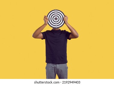 Unknown Dark-skinned Man In Casual Clothes Hides His Face Behind Darts Board On Yellow Background. Concept Of Defining Business Goals, Finding Target Audience, Achieving Goals And Success.