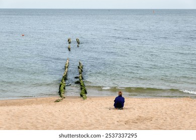 An unknown elderly woman sits on the beach near an old breakwater and watches the sea waves rolling onto the shore. Background. Landscape. - Powered by Shutterstock