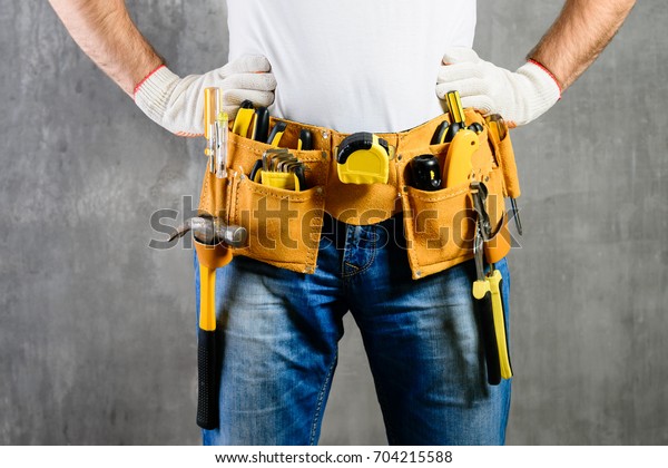 unknown handyman with hands on waist and tool\
belt with construction tools against grey background. DIY tools and\
manual work concept