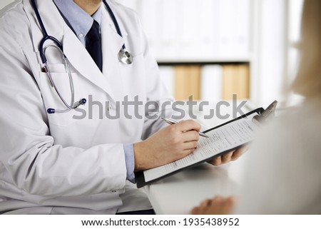 Unknown male doctor and patient woman discussing current health examination while sitting in clinic and using clipboard. Perfect medical service in hospital. Medicine and healthcare concept