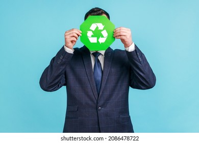 Unknown Male Person Wearing Official Style Suit Hiding Face Behind Green Recycling Sign, Garbage Sorting And Environment Protection, Thinking Green. Indoor Studio Shot Isolated On Blue Background.