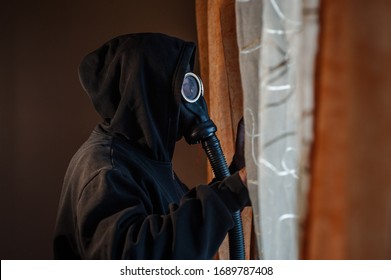 Unknown Man Wearing Black Hood And Protective Gas Mask Standing By The Window At Home Looking Trough The Window Side View