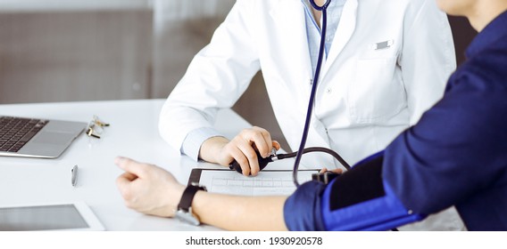 Unknown Woman Doctor Is Checking Her Patient's Blood Pressure While Sitting Together In Clinic. Medicine Concept During Coronavirus Pandemic. Covid 2019