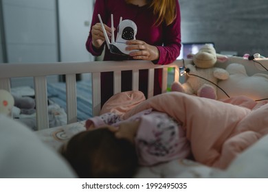 Unknown Woman Mother Adjusting And Setting Up Surveillance Security Camera On Baby Bed At Home In Bedroom Watching Small Child In Cradle While Sleeping In Dark Room At Night