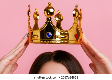 Unknown woman putting on golden crown, arrogance and privileged status, concept of self confidence in success, self-motivation and dreams to be best. Indoor studio shot isolated on pink background.