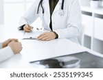 Unknown woman-doctor and female patient sitting and talking at medical examination in clinic, close-up. Therapist wearing green blouse is filling up medication history record. Medicine concept