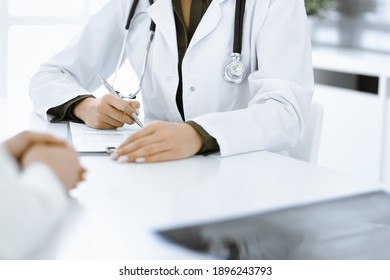 Unknown woman-doctor and female patient sitting and talking at medical examination in clinic, close-up. Therapist wearing green blouse is filling up medication history record. Medicine concept