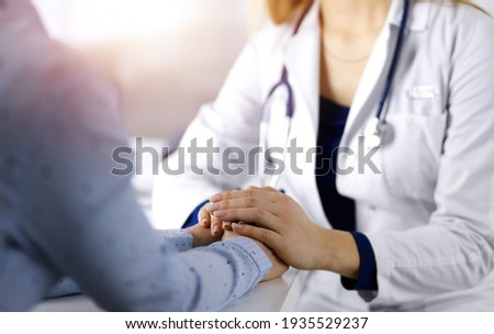 Unknown woman-doctor is holding her patient's hands to reassure the patient, discussing current health examination, while they are sitting together at the desk in the sunny cabinet in a clinic. Female
