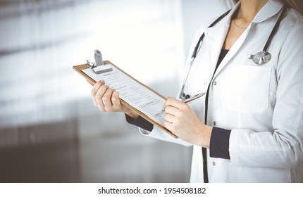 Unknown woman-doctor is writing some notes using a clipboard, while standing in the cabinet in a clinic. Female physician with a stethoscope, close-up. Perfect medical service in a hospital. Medicine