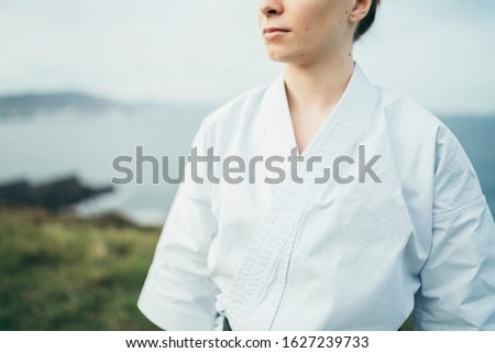 Unknown young female karate athlete looking away. She is wearing white kimono. She is outdoor on top of a cliff. She is redhead and freckles. Blue sky and sea with bokeh effect on background.
