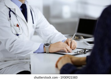 Unknown young woman patient discuss the results of her medical tests with a doctor, while sitting at the desk in a hospital office. Physician using clipboard for filling up medication history records