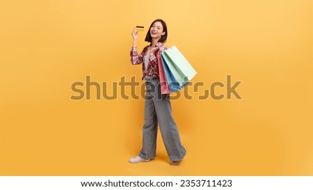 Unlimited shopping concept. Asian lady holding colorful shopper bags with gifts and showing credit card over yellow studio background, panorama with copy space, full length