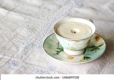 An Unlit Soy Candle In A Fine China Teacup On A White Linen With Blue Embroidery