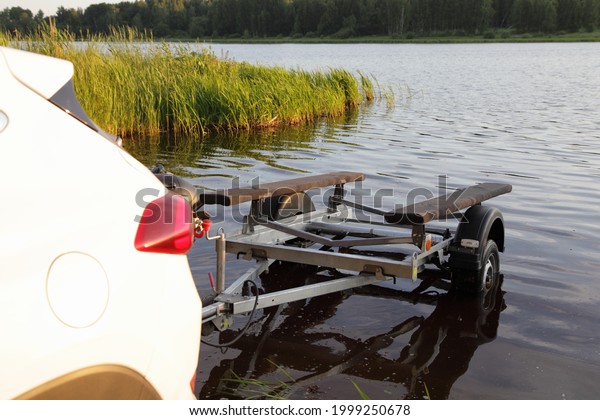 Unloaded empty boat\
trailer on sandy shipway on the river water and new white SUV\
crossover car, boat launch before active recreation on the\
countryside river in\
Europe
