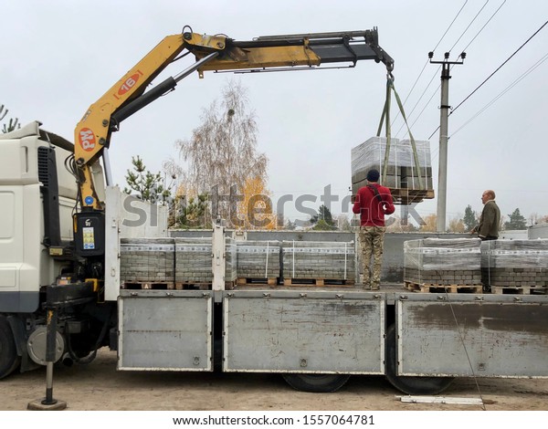 Unloading paving slabs from a truck. Men unload\
paving slabs using a manipulator. Workers unload building materials\
from a large machine. Dmitrovka, Kiev region, November 8,\
2019.
