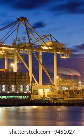 The Unloaing Of A Cargo Vessel Using Huge Overhead Cranes And Carts, Transporting The Containers To And From The Ship. Some Smoke Stacks In The Distance