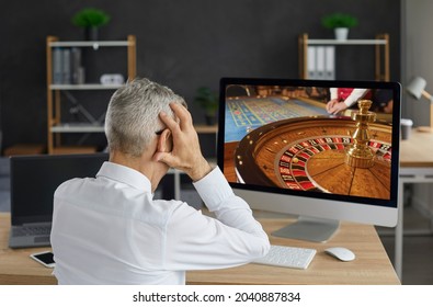 Unlucky Old Gambler Places Bet In Online Casino Gambling Game And Loses All His Money. Back View Of Senior Man Looking At Desktop Computer PC Screen Feeling Desperate And Holding His Head In His Hands