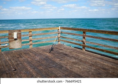 Unmanned Fishing Rod On A Wooden Dock Or Pier Balancing On The Rails Overlooking A Calm Blue Ocean On A Sunny Day