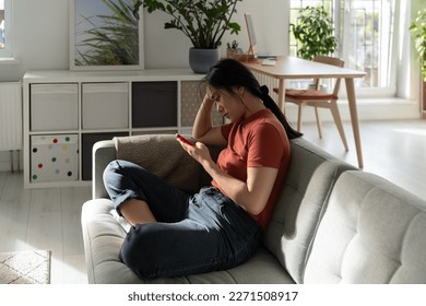 Unmotivated confused Asian woman sits with phone on sofa in apartment not willing to start working. Procrastinating Chinese student girl is addicted to mobile gadgets and Internet surfing 