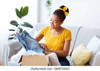 Unpacking Clothes. Portrait of excited afro lady opening and unboxing parcel cardboard box or putting jeans in, sitting on sofa in living room at home. Young woman satisfied with delivery service