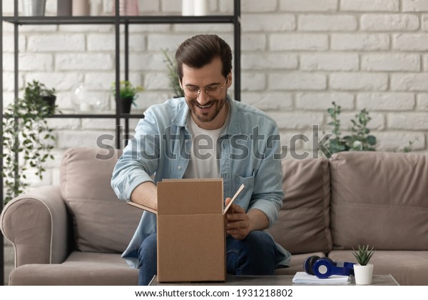 Unpacking parcel. Happy young man postal delivery\
service client sit on couch at living room open small carton box\
enjoy goods received. Curious guy online shopper get new order from\
web store by mail