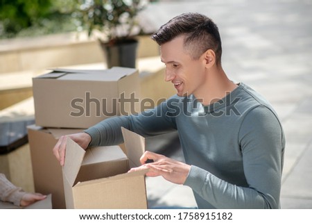 Unpacking. Smiling handsome man sitting next to the cardboards and opening one