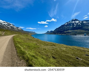 unpaved road next to the blue water of an Icelandic fjord surrounded by snow capped mountains on a sunny day - Powered by Shutterstock