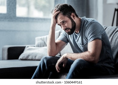 Unpleasant pain. Sad unhappy handsome man sitting on the sofa and holding his forehead while having headache