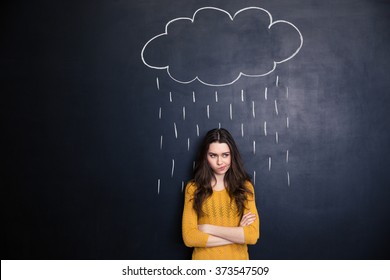 Unpleased young woman with raincloud drawn over her on a blackboard background standing with arms crossed 