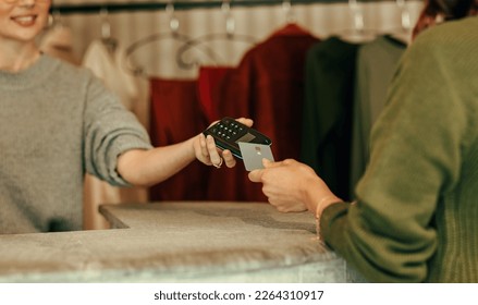 Unrecognisable female customer paying with a credit card at the checkout counter. Young woman tapping a credit card on a contactless card reader. Woman purchasing clothes in a clothing store. - Shutterstock ID 2264310917