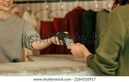 Unrecognisable female customer paying with a credit card at the checkout counter. Young woman tapping a credit card on a contactless card reader. Woman purchasing clothes in a clothing store.