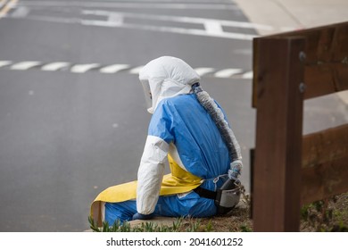 An Unrecognisable Male Medic Sitting Outside The Hospital In A Bio Weapon Protective Suit In A Daytime, Resting On The Bench