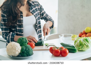 Unrecognisable pregnan woman cook salad cutting cucumber on wooden cutting board putting fresh vegetables and fruit on table in kitchen. Pregnancy, maternity, healthy lifestyle concept.