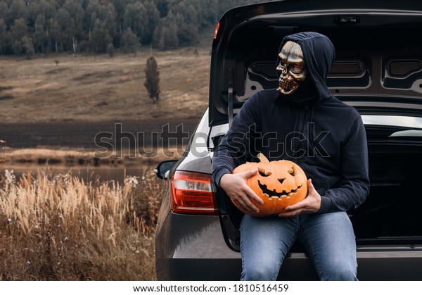 An unrecognizable adult man wearing a skull\
mask sits in the trunk of a car with carved pumpkins for Halloween,\
outdoors. Trick or trunk. Copy\
space.