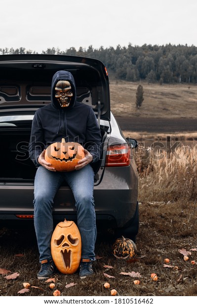 An unrecognizable\
adult man wearing a skull mask sits in the trunk of a car with\
carved pumpkins for Halloween. Trick or trunk. Vertical\
orientation, copy space,\
outdoors.