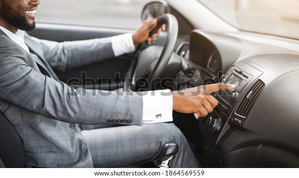 Unrecognizable african american man driver turning\
on music in car before moving. Black businessman pushing button on\
functional panel in his car, turning on music, panorama with empty\
space
