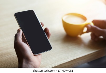 Unrecognizable African American Man Holding Smartphone With Empty Screen And Cup Of Coffee At Table In Cafeteria, Closeup. Mockup For Design