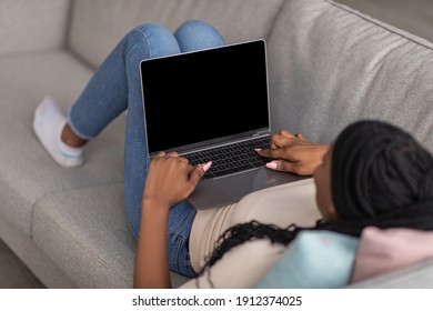 Unrecognizable African American Woman Laying On Sofa At Home And Using Modern Laptop With Blank Screen, Mockup, Copy Space. Black Lady Typing On Computer Keyboard, Empty Display