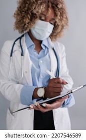 Unrecognizable African Ethnicity Doctor With Curly Hair In A Medical Mask In A Lab Coat, Writes Information To The History Of The Disease On A Clipboard. Concept: Medicine, Doctor, First Aid