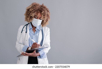 Unrecognizable African Ethnicity Doctor With Curly Hair In A Medical Mask In A Lab Coat, Writes Information To The History Of The Disease On A Clipboard, Isolated On Grey Background Mocap 