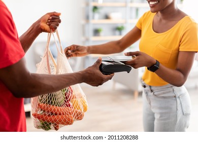 Unrecognizable African Lady Paying Via Cellphone For Grocery Shopping Delivery, Receiving Food Products From Courier Guy Standing At Home. Cropped Shot, Selective Focus On Phone