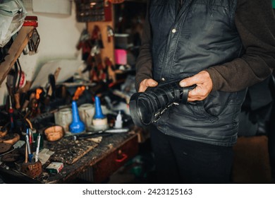 An unrecognizable artisan holding boots for repair while standing at shoemaker workshop. Cropped picture of an old shoemaker holding boots in his shop and preparing for repairing. Small business. - Powered by Shutterstock