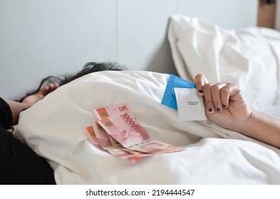 Unrecognizable Asian Woman Hands Holding Condom And Indonesian Rupiah Cash On A Bed Under The Blanket. Focus At Hand. Prostitution Concept.