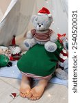 an unrecognizable barefoot child on the floor, a soft toy teddy bear sitting on his back. Cozy homely holiday atmosphere. holidays at home, pampering and fun on Christmas Eve