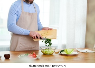 Unrecognizable Bearded Host Of Cooking TV Show Standing At Wooden Table And Preparing Vegetable Salad, Interior Of Modern Studio With Panoramic Windows On Background